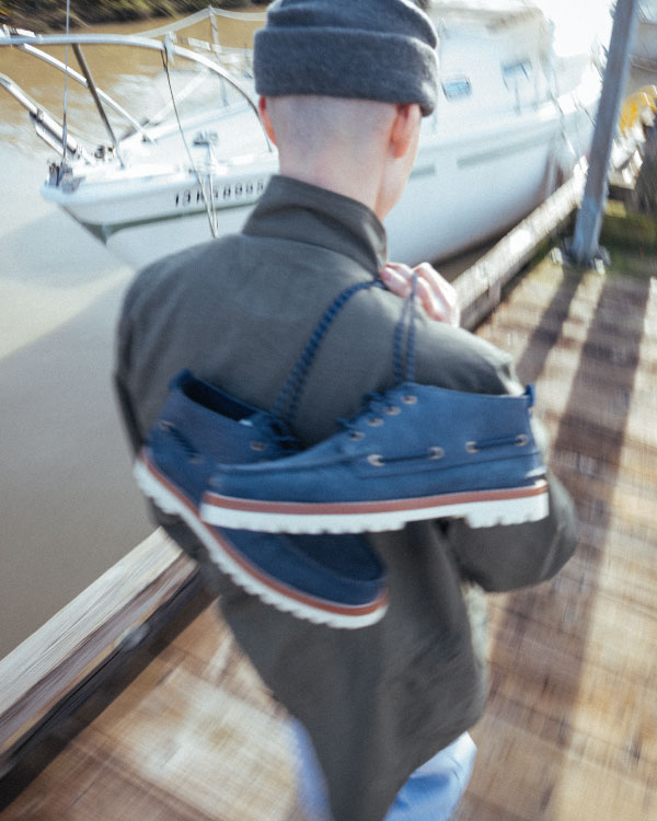 A person walking along a dock beside a sailboat with the Herschel Supply Company Authentic Original Lug Chukka Sperry shoes in Peacoat/Navy/Blanc de Blanc