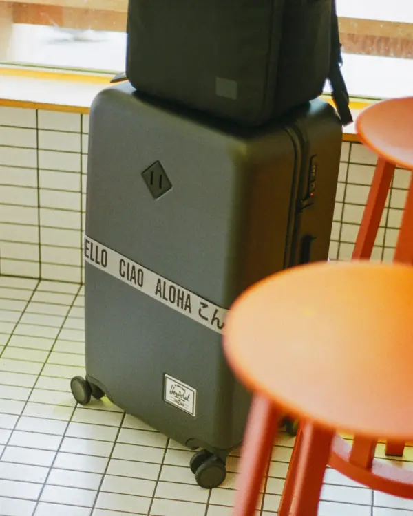 a grey large herschel heritage hardshell luggage with a luggage belt sits in the corner of a cafe 