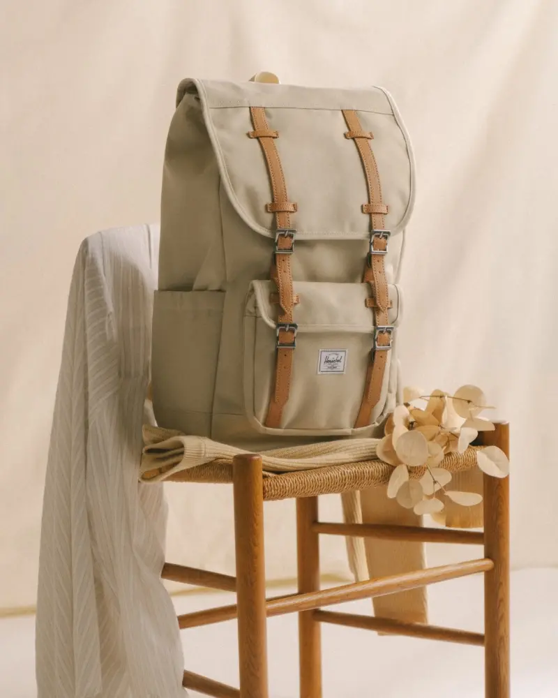 A photo of a Herschel Little America® Backpack in the new seasonal color, Eucalyptus, on a wooden chair with a soft beige backdrop and some eucalyptus leaves on the chair beside it.